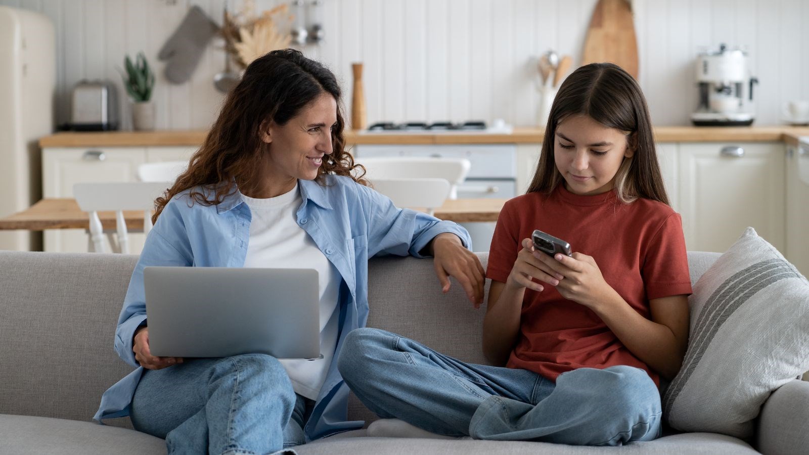 Mom with a laptop and daughter on a cell phone sit together on a couch.
